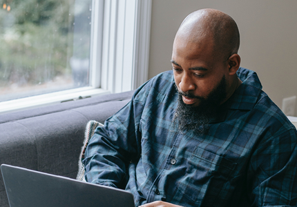 Black man sitting on sofa next to a window working on his laptop